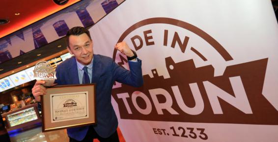 Zdjęcie zrobione podczas gali Made in Toruń. Laureat stoi na tle grafiki Made in Toruń. Prawą dłonią trzyma certyfikat, lewą zaciska i unosi do góry.